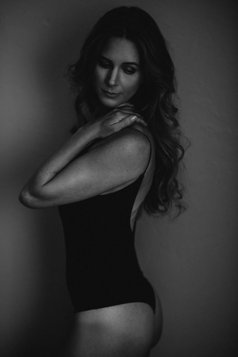 Black and white portrait of a woman with long wavy hair, wearing a dark sleeveless top, looking down with a contemplative expression, arms crossed near her shoulders.