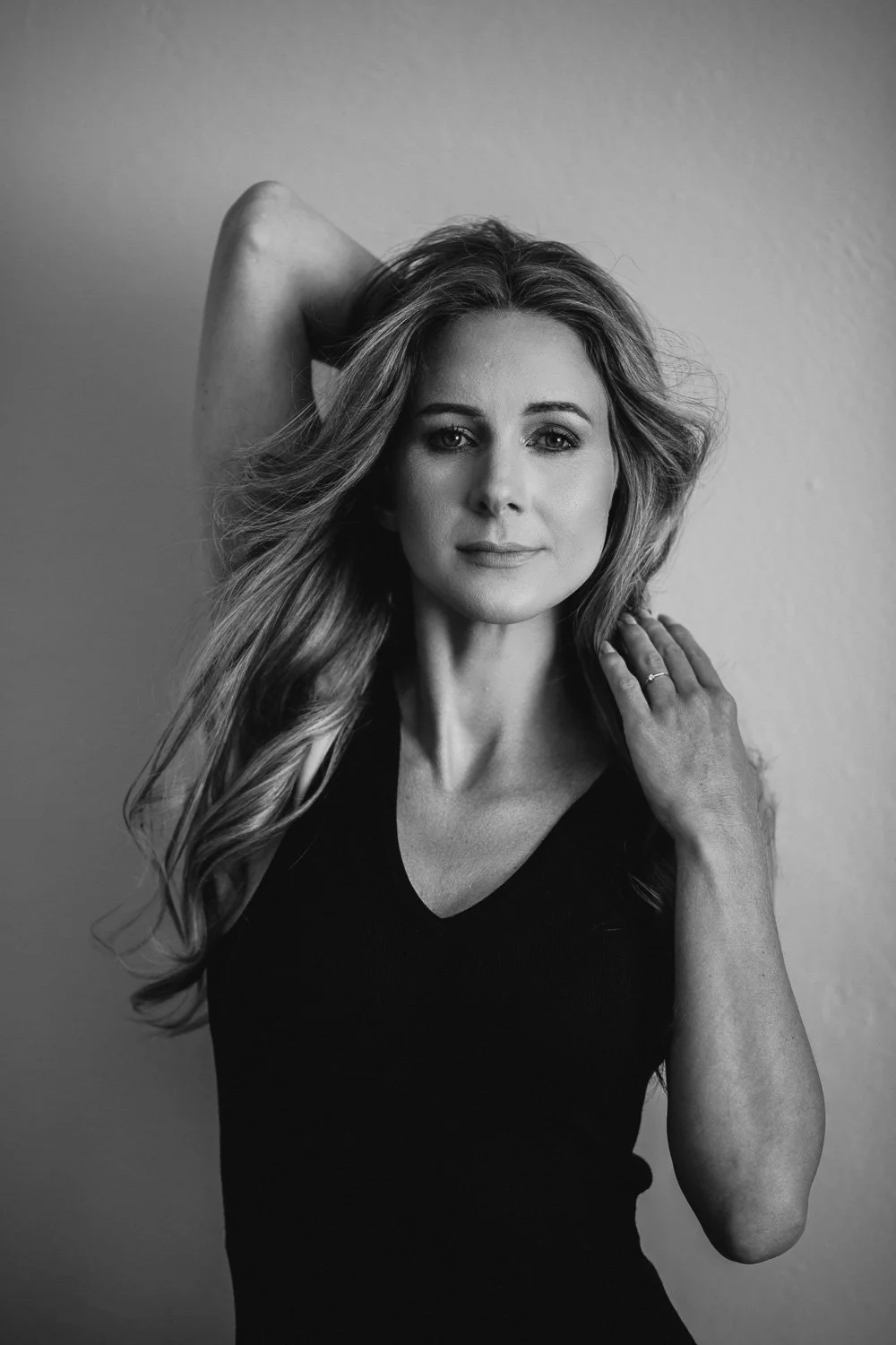 Black and white portrait of a woman with long, wavy hair, wearing a black top, looking at the camera with one arm raised and her hand behind her head, and the other hand resting on her shoulder.