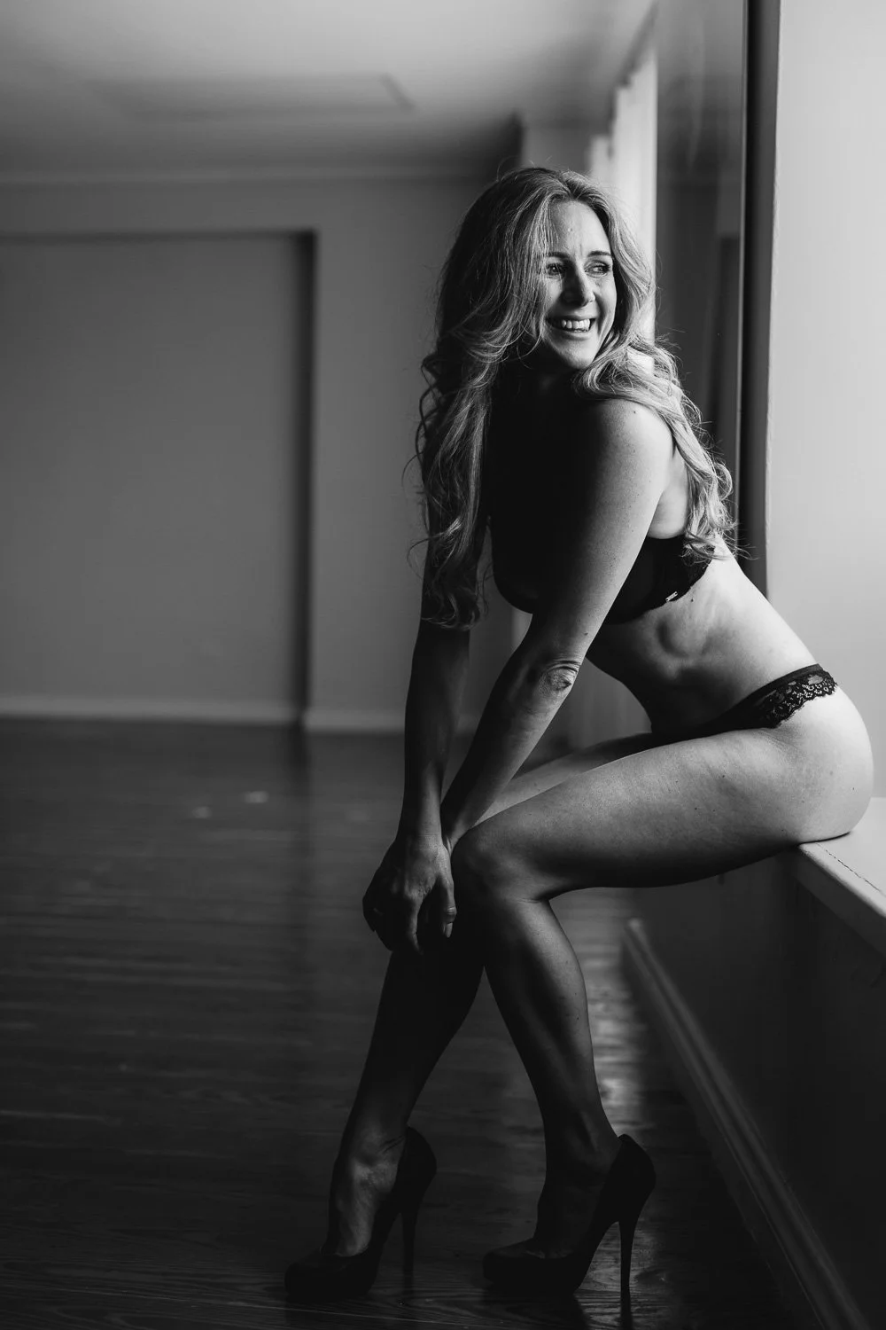 A woman wearing lingerie and high heels sitting by a window, smiling and looking out.