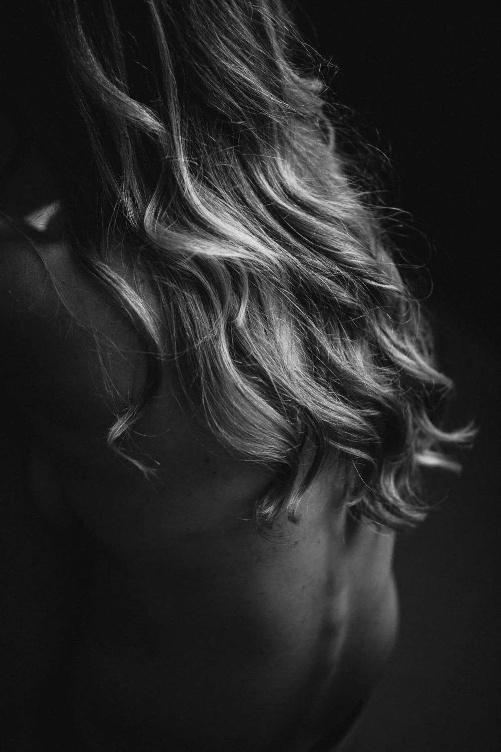 Black and white close-up of wavy, shoulder-length hair on a person, with part of their shoulder and neck visible.