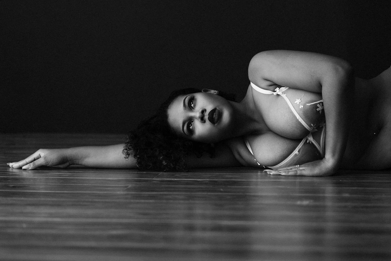 Black and white photo of a woman lying on the floor on her side with her head resting on her arm, wearing a floral lingerie top, with curly hair and bold makeup.