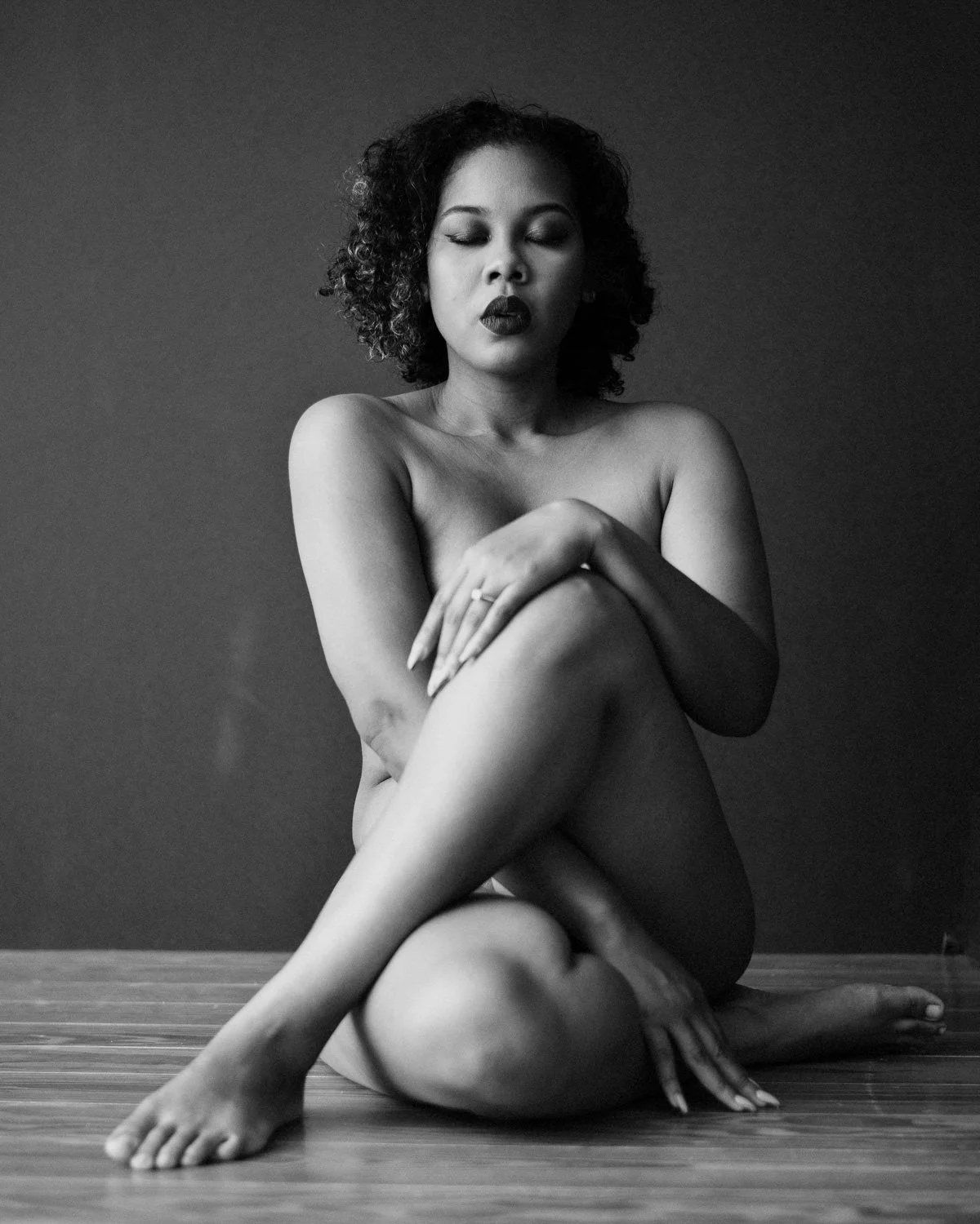 A black and white photo of a woman with curly hair sitting on the floor with her legs crossed and arms folded over her knees, eyes closed, with a calm expression.