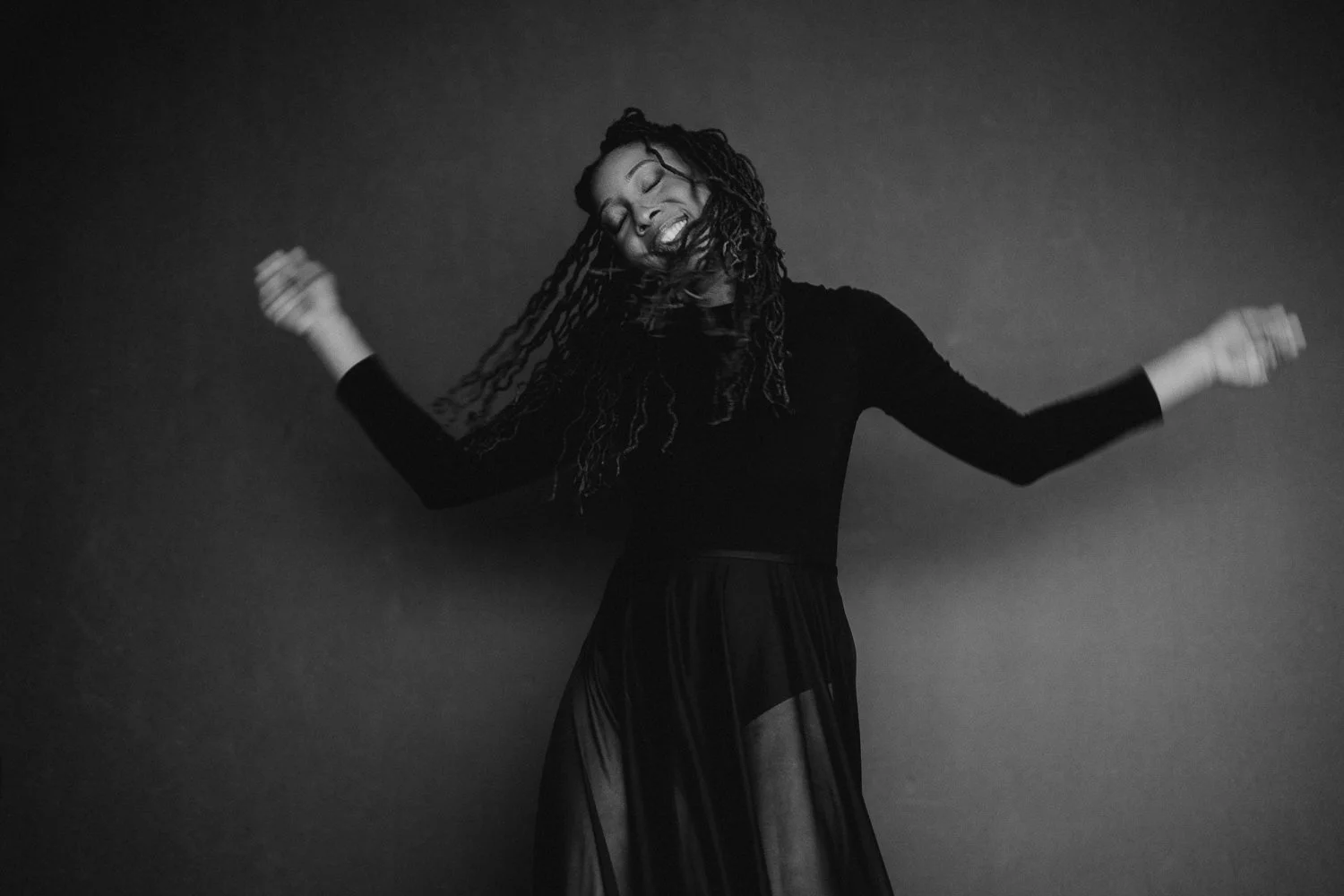 Black and white photo of a woman with long dreadlocks dancing with a big smile on her face, wearing a long sheer skirt and a long sleeve top.
