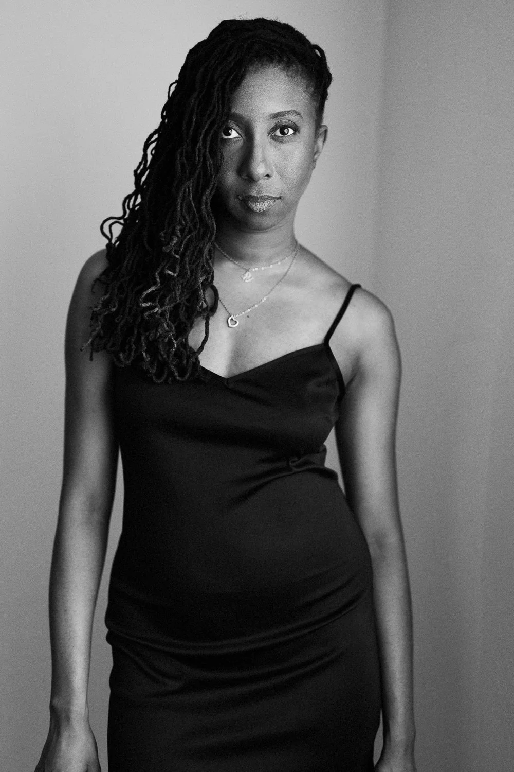 A woman with dreadlocks, wearing a black spaghetti strap dress and necklaces, standing against a plain wall in a black and white photograph.