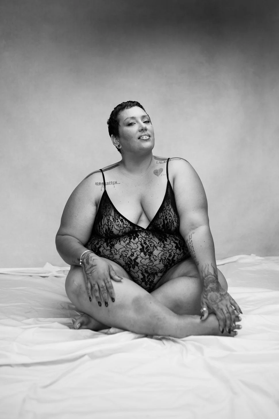 A woman with short dark hair, tattoos, and pierced nose, sitting cross-legged on a bed in a black lace lingerie top, in a black and white photo.