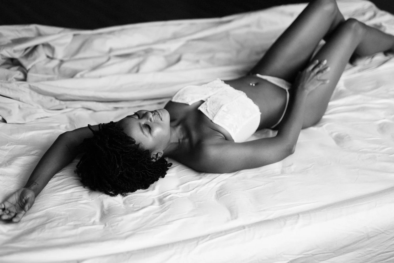 A woman with dark skin and curly hair lying on a bed with white sheets, wearing a strapless top and underwear, with her eyes closed.