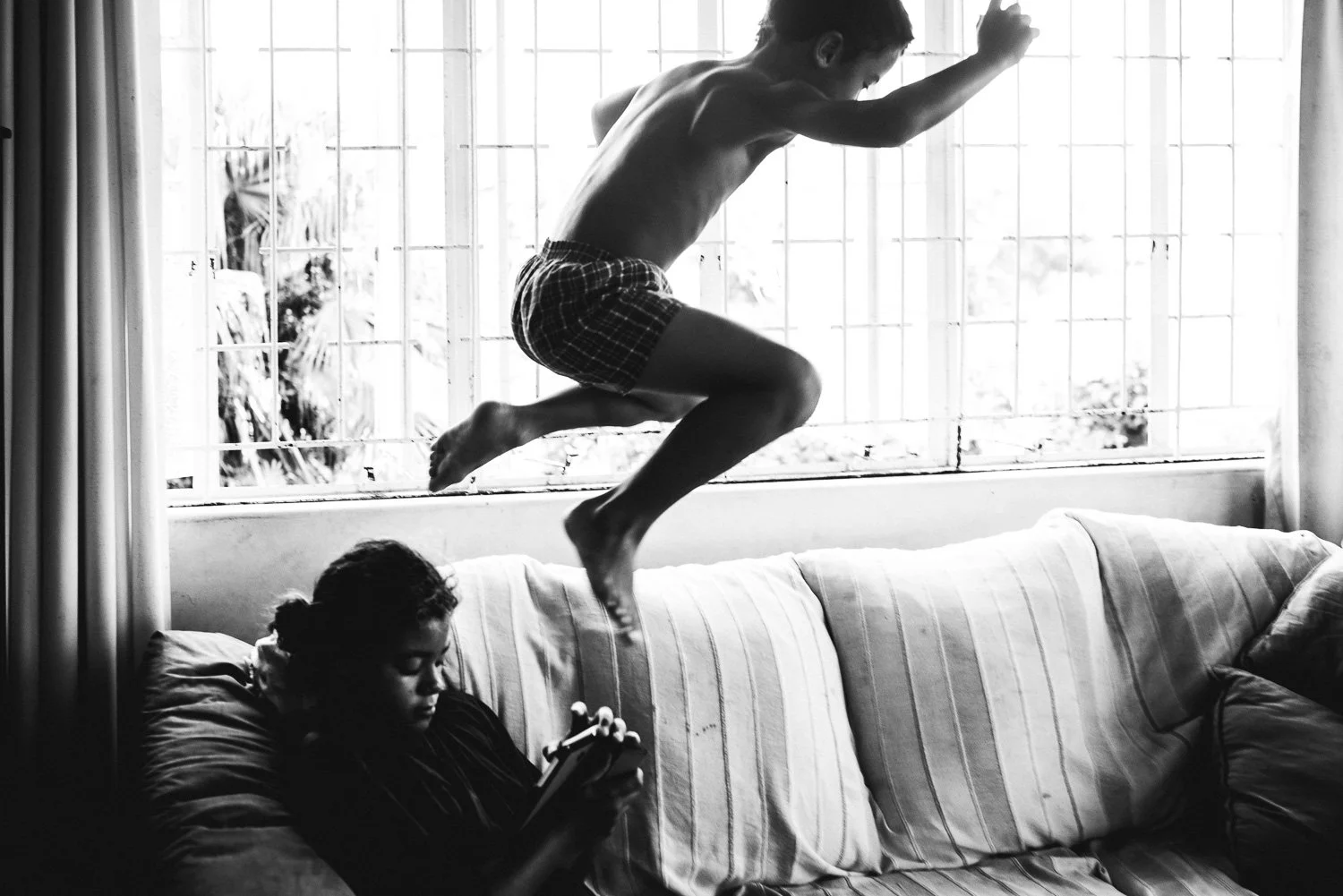 A black and white photo of a boy jumping onto a sofa while a girl sits on the sofa below, looking at her phone in a room with large windowpanes.