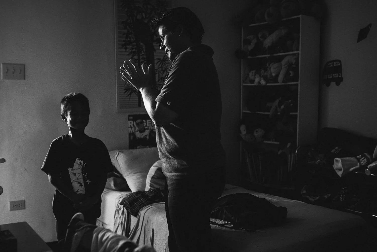 A black and white photo of an adult and a child indoors. The adult is standing with hands pressed together in a prayer-like gesture, facing the child who is standing and smiling. The background shows a bed, a window with blinds, and a bookshelf fille