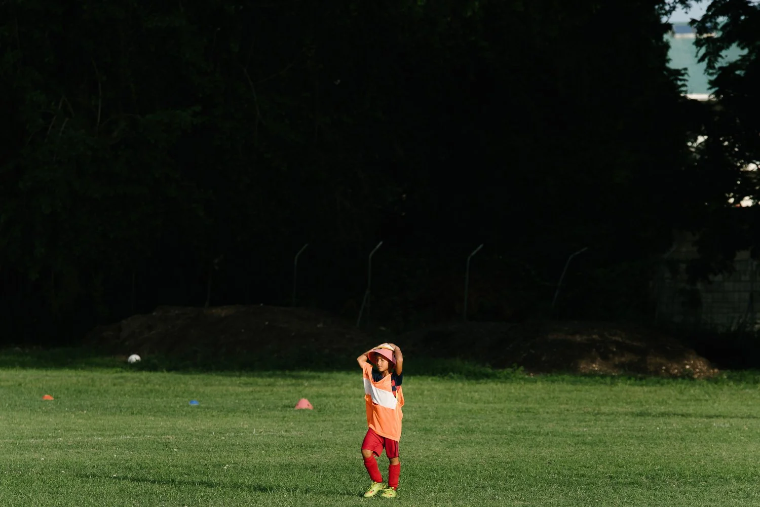 A young boy in a soccer uniform standing on a grassy field, wearing a pink hat and holding his head with both hands, with colorful cones and trees in the background.