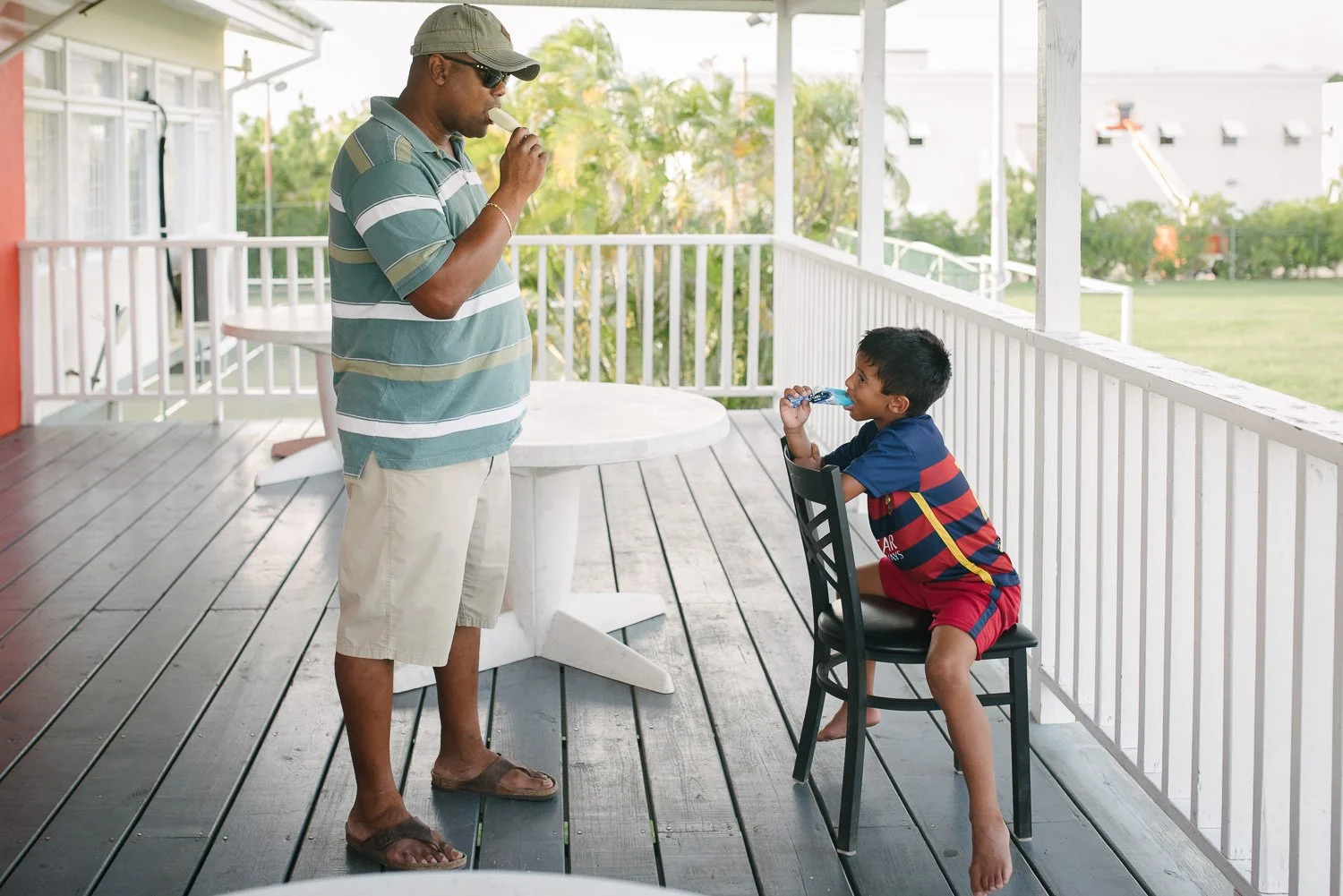 A man and a boy on a porch with white railings, with the man standing and the boy sitting on a black chair, both holding and eating ice cream cones on a sunny day.
