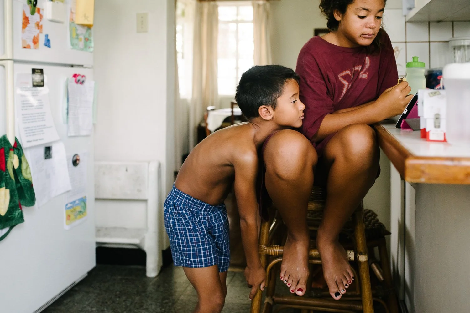 A young boy in plaid shorts peeks over a chair to look at a girl at the kitchen counter using a tablet.