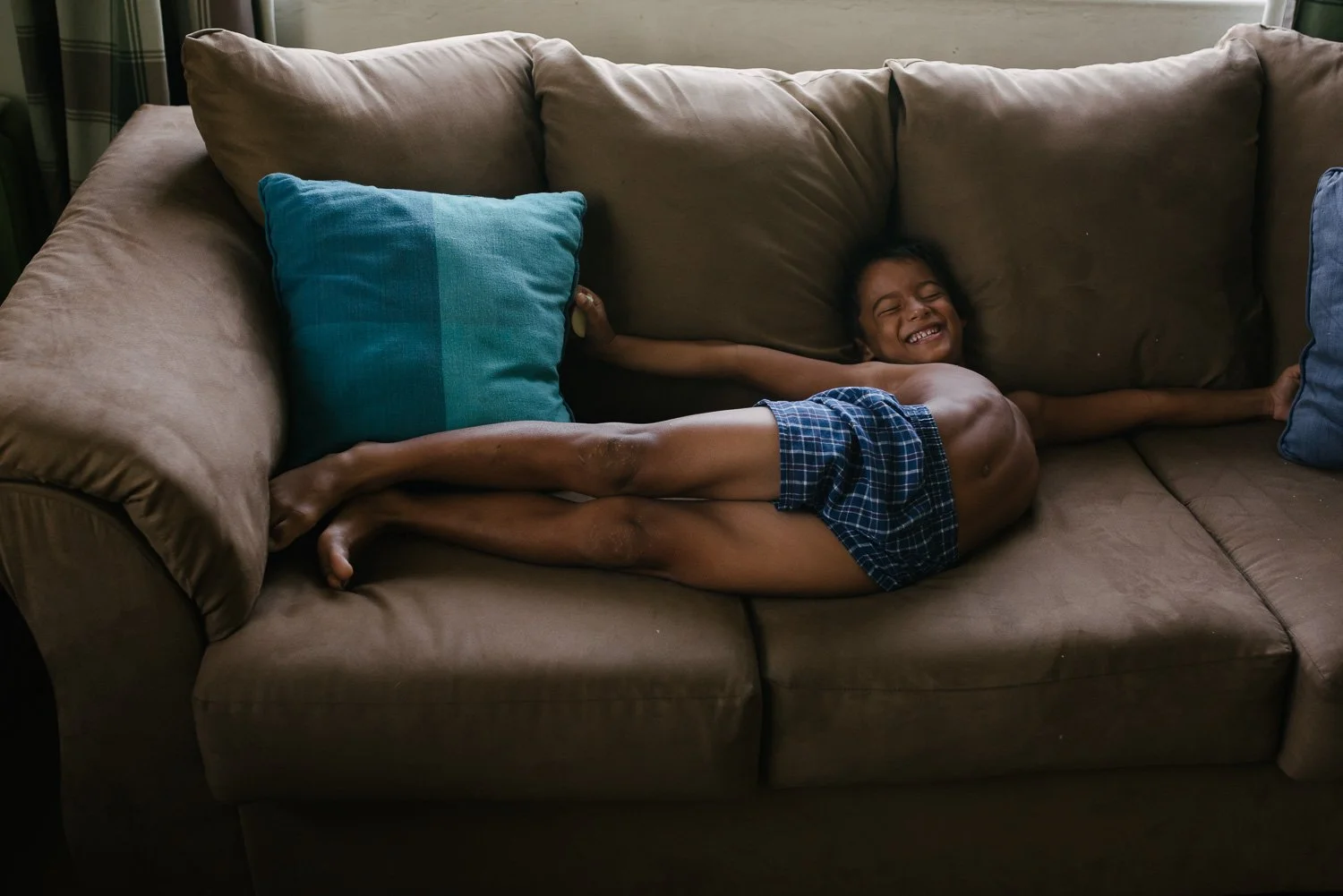 A young boy lying on a brown couch, smiling with eyes closed, wearing blue plaid shorts, with a large turquoise pillow behind his back and a smaller dark blue pillow near his feet.
