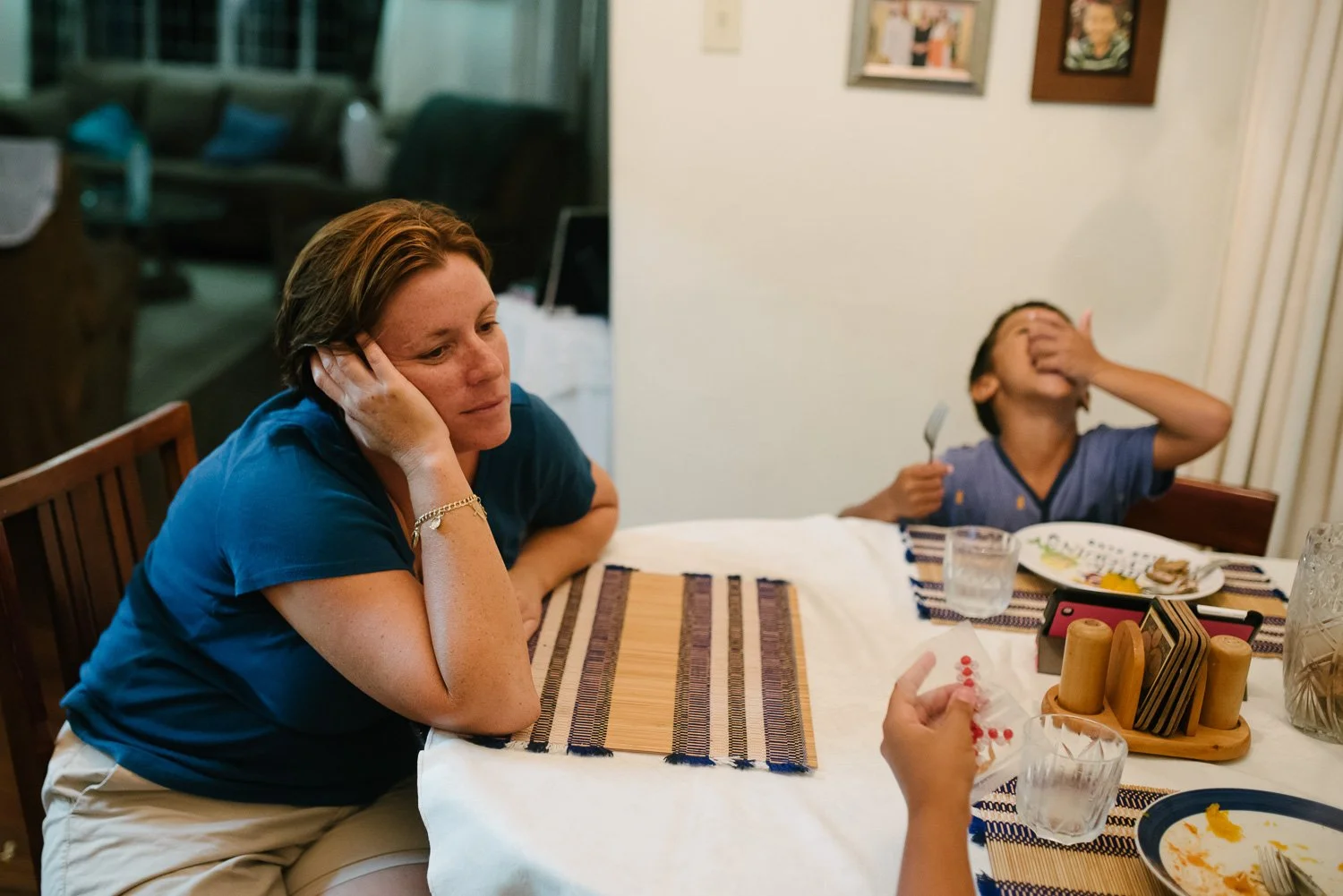 Woman sitting at a dining table with her hand on her face, looking down, while a young boy in the background is covering his face with his hand and holding a fork, with food and glasses on the table.