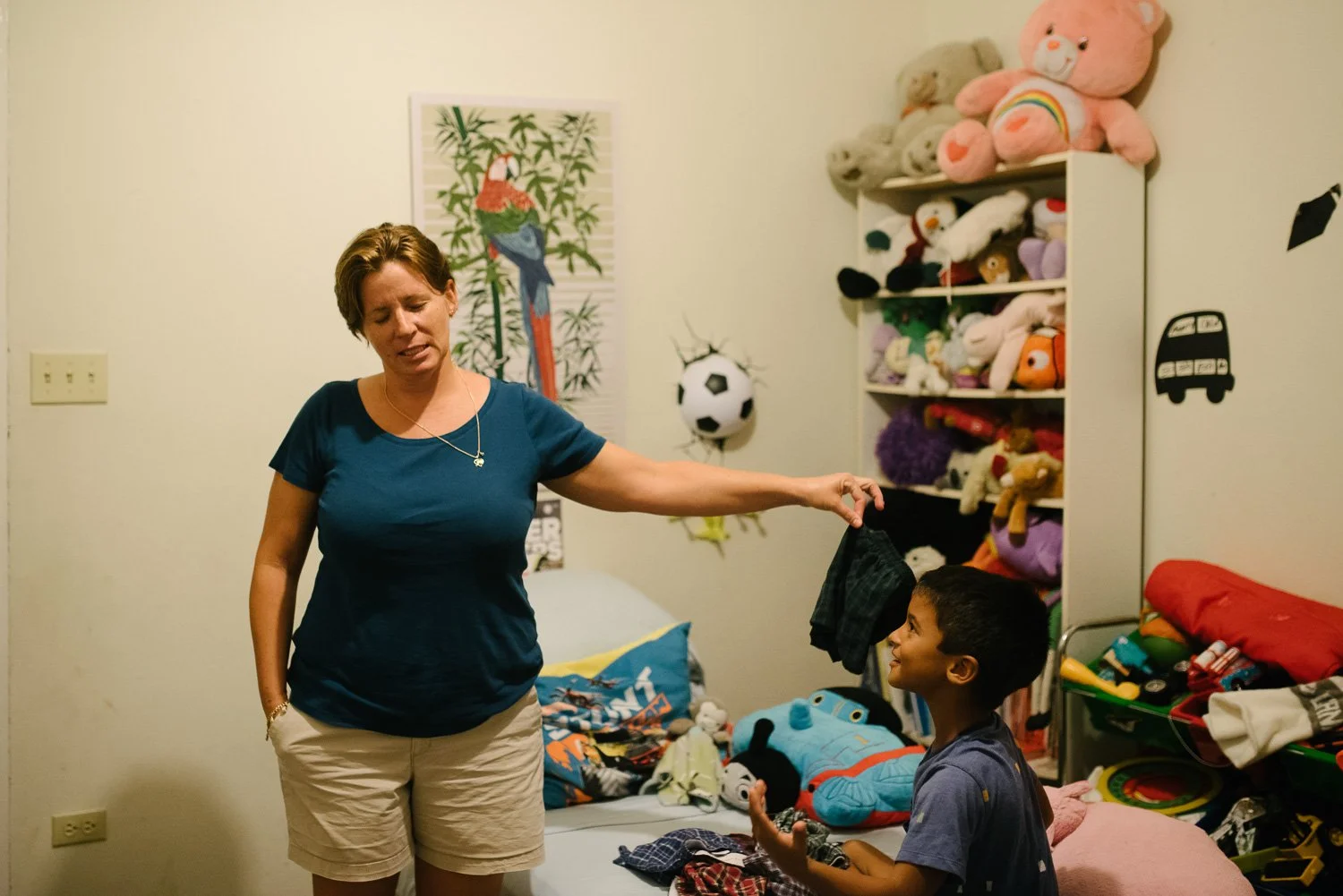 A woman and a boy in a bedroom with toys and stuffed animals, woman holding clothing and the boy smiling.