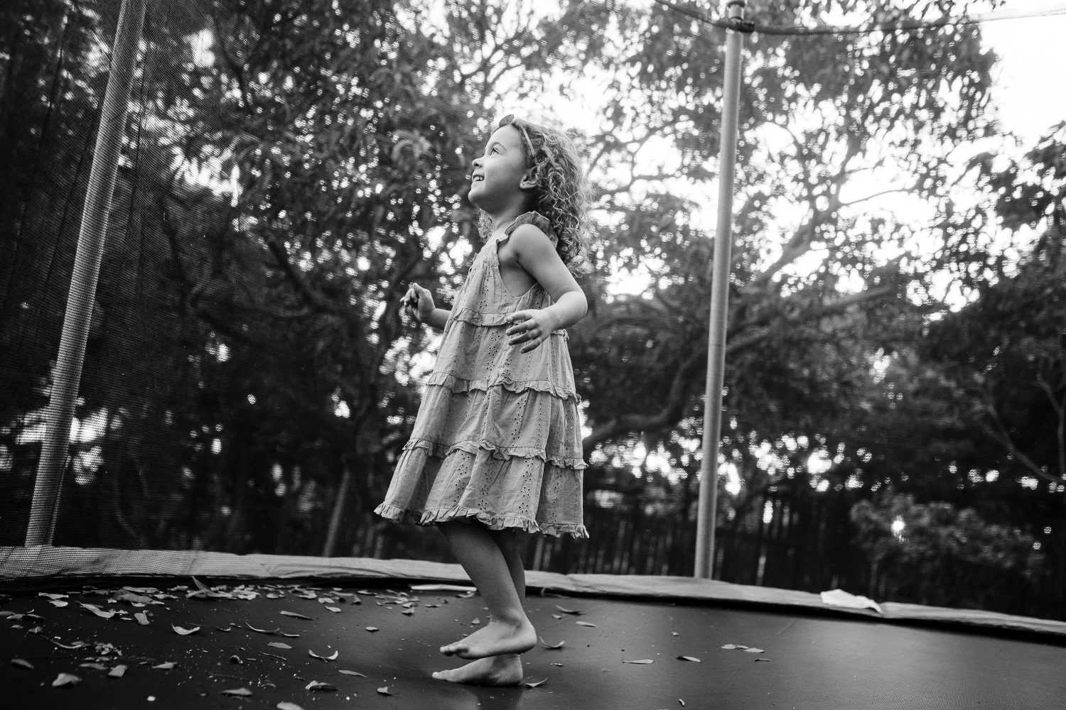 A young girl with curly hair, wearing a tiered dress, is jumping on a trampoline outdoors surrounded by trees.