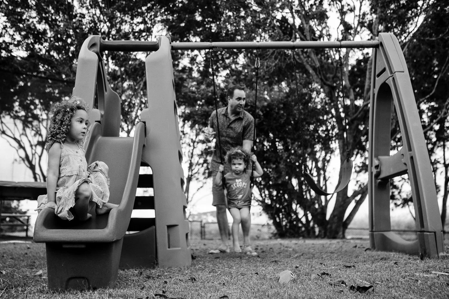A black and white photo of a family in a park, with two children on swings, and an adult pushing one of the children.