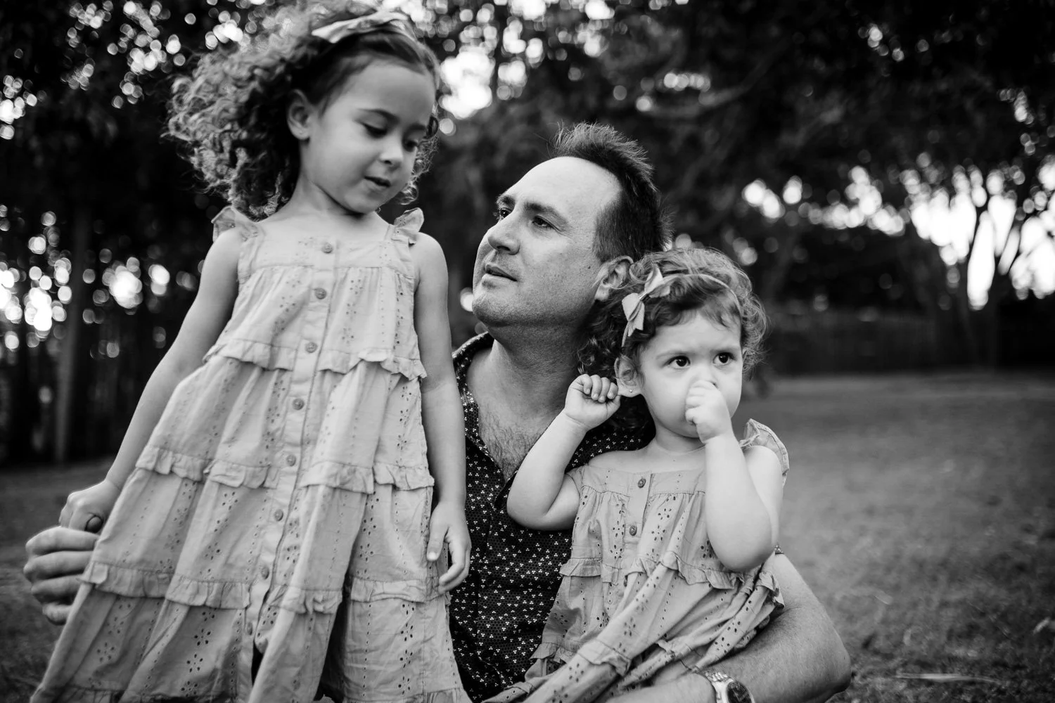 A man outdoors with two young girls, one standing and the other sitting, both wearing dresses, in black and white.