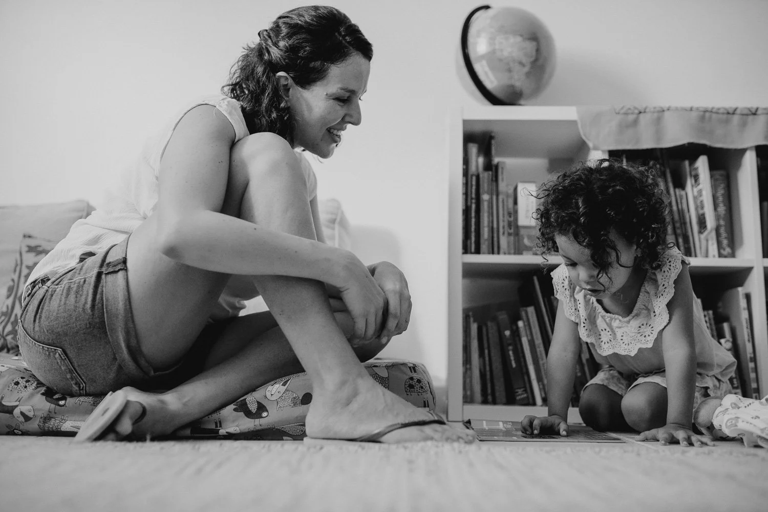 A woman and a young girl are sitting on the floor, engaging in play or reading together. The woman is sitting cross-legged, smiling, while the girl is focused on a book or a game on the floor. There is a bookshelf with books in the background.