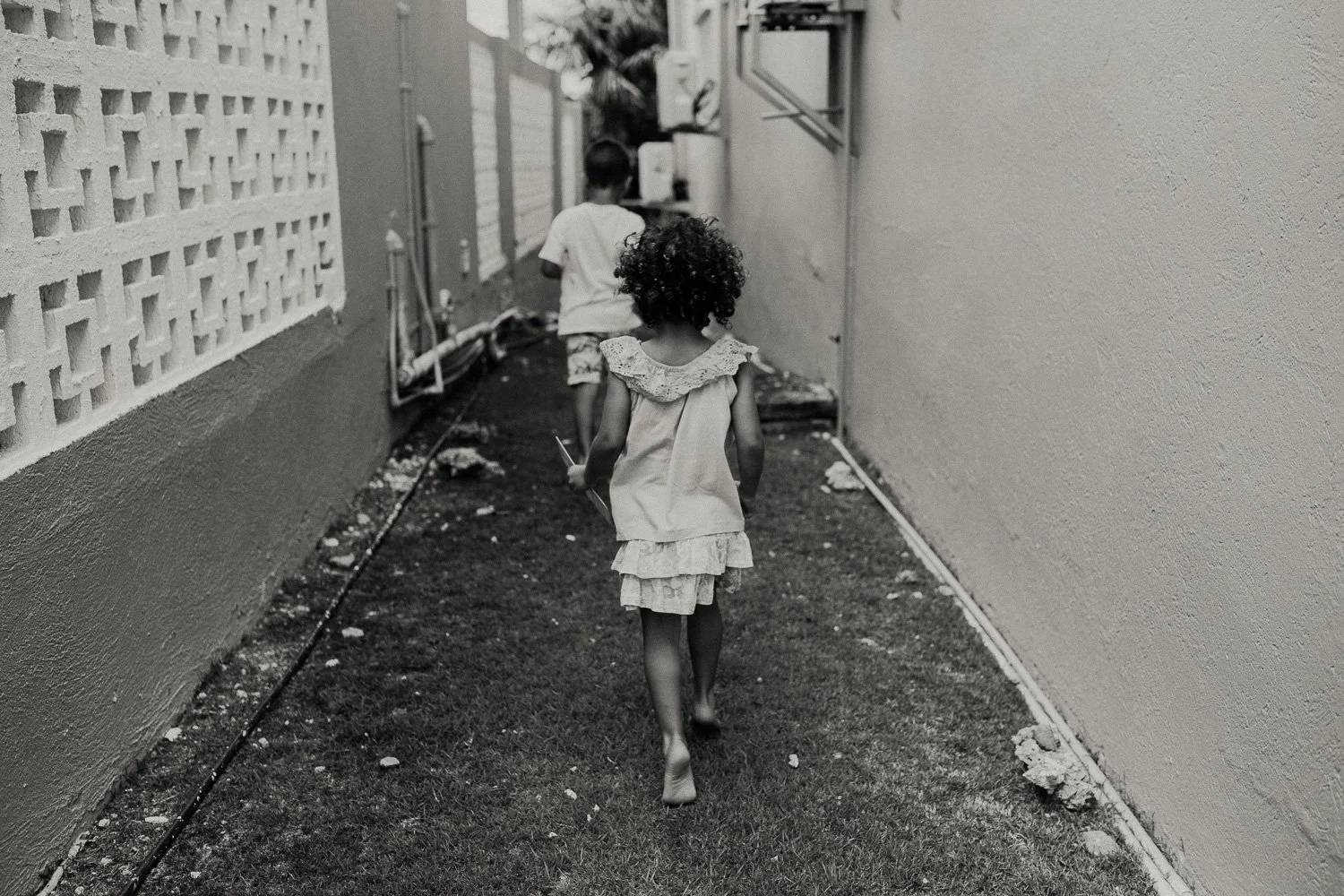 Two young children walking down a narrow outdoor alley between two walls, one girl with curly hair in the foreground and a boy with straight hair and shorts in the background, surrounded by small debris.