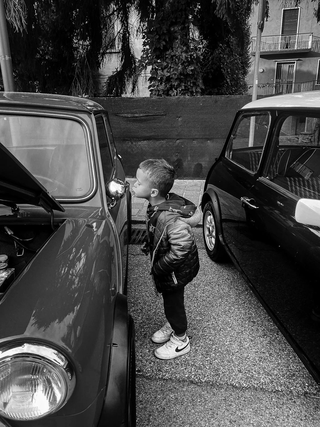 A young boy in a jacket and sneakers is looking closely at the side mirror of a vintage car parked between two other cars on a city street.