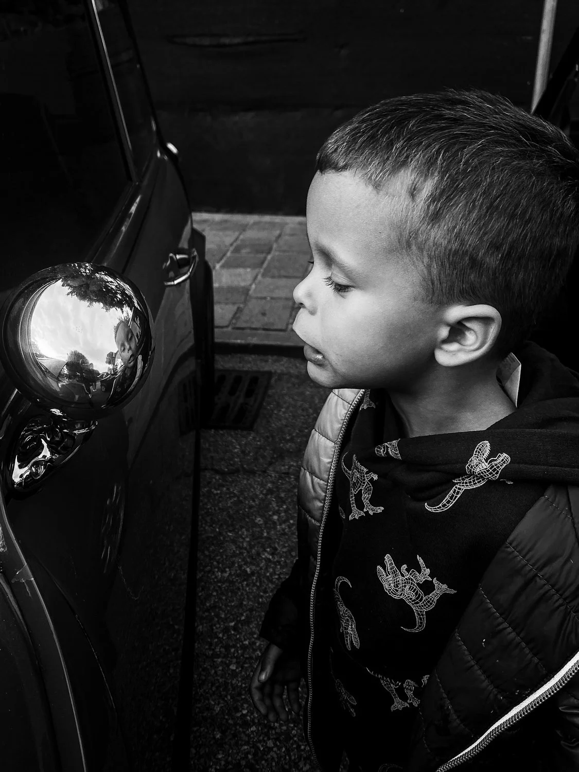 A young boy with short hair, eyes closed, leaning towards a shiny, rounded vehicle side mirror, reflecting trees and the sky.