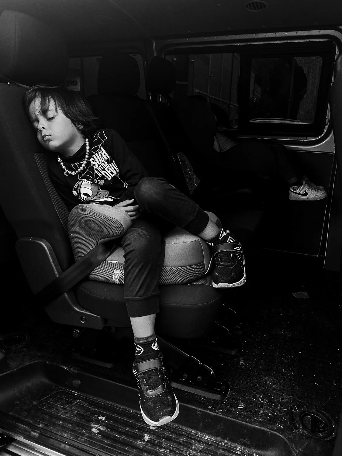 A young boy sleeping in a car seat on a vehicle's bus or van, with another person sleeping nearby. The boy is wearing a graphic t-shirt, sneakers, and a beaded necklace.