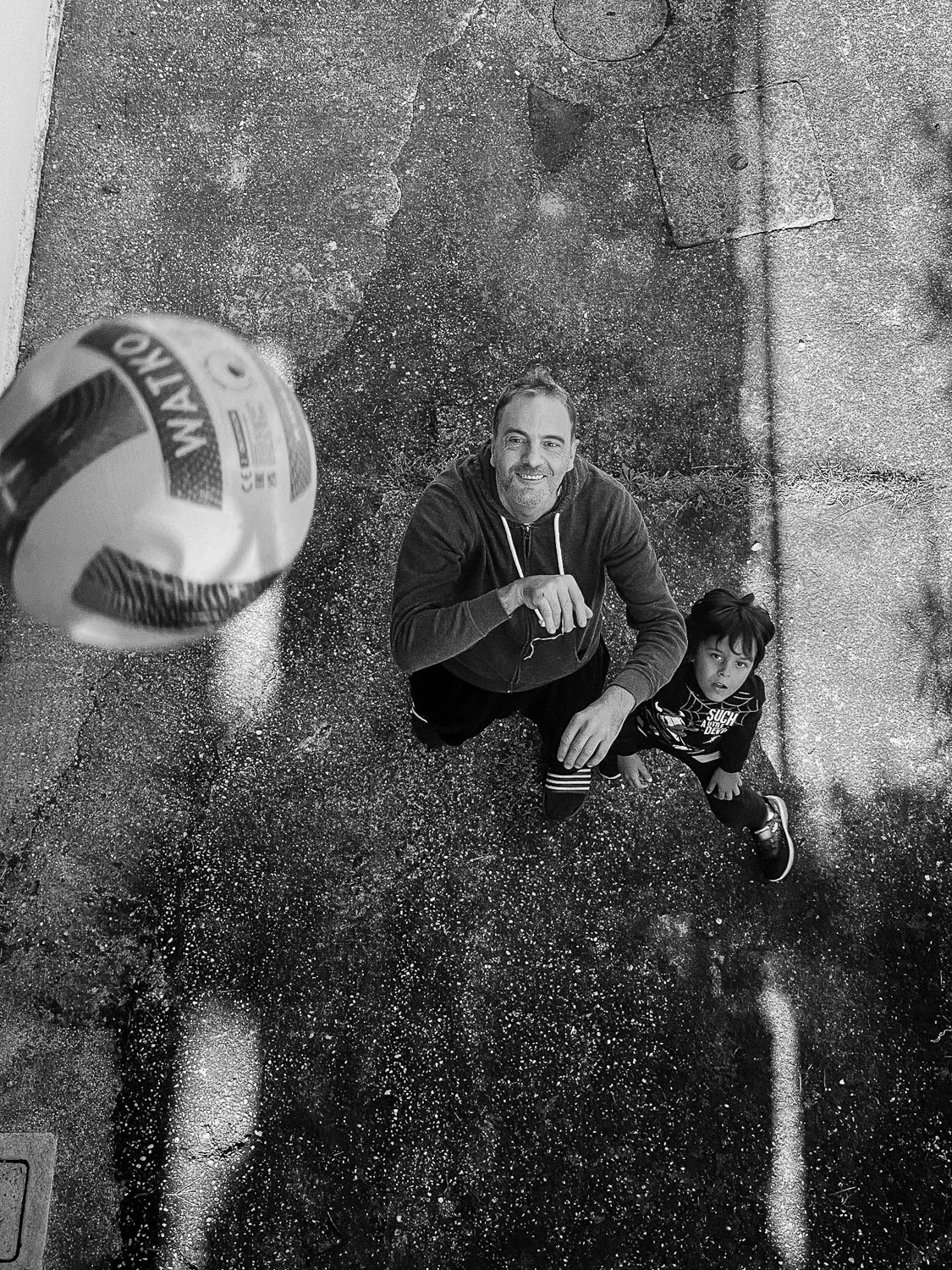 A man and a young boy looking up at a ball in the air, taken from above at ground level on a rough, textured outdoor surface.