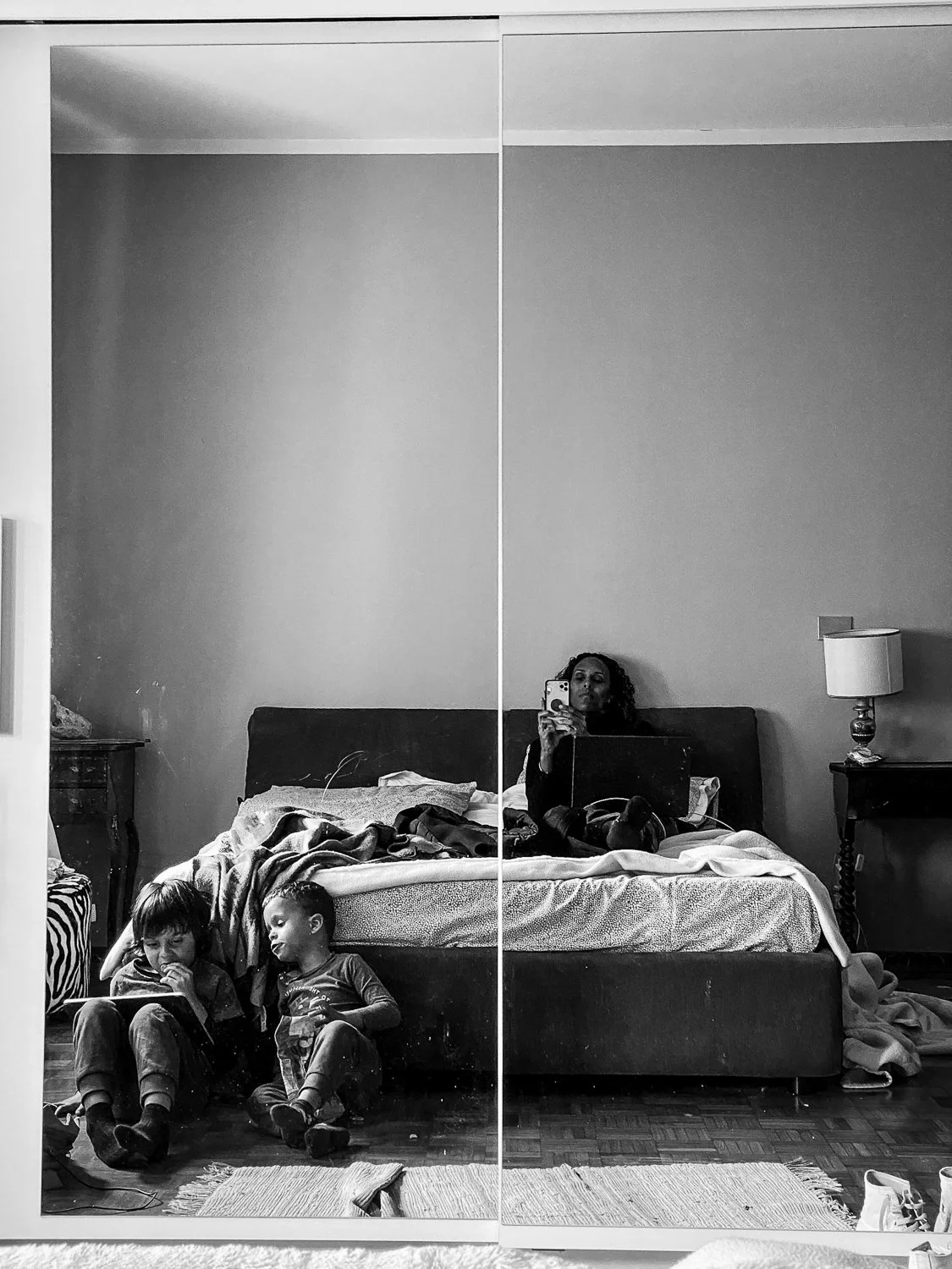 A black and white mirror selfie of a woman taking a photo, with two children sitting on the floor in front of her, all in a bedroom with a bed, bedside tables, and a lamp.