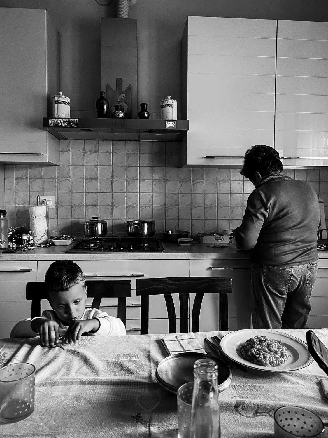 A black and white photo of a kitchen with two people, a child at a dining table and an adult cooking or preparing food in the background. The child is sitting at the table with a focused expression, reaching toward the table. The table has a large pl