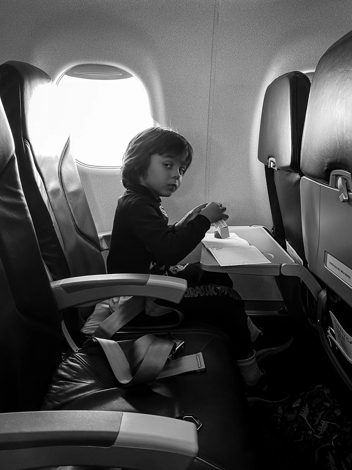A child sitting in an airplane seat, looking towards the camera, with a window in the background.