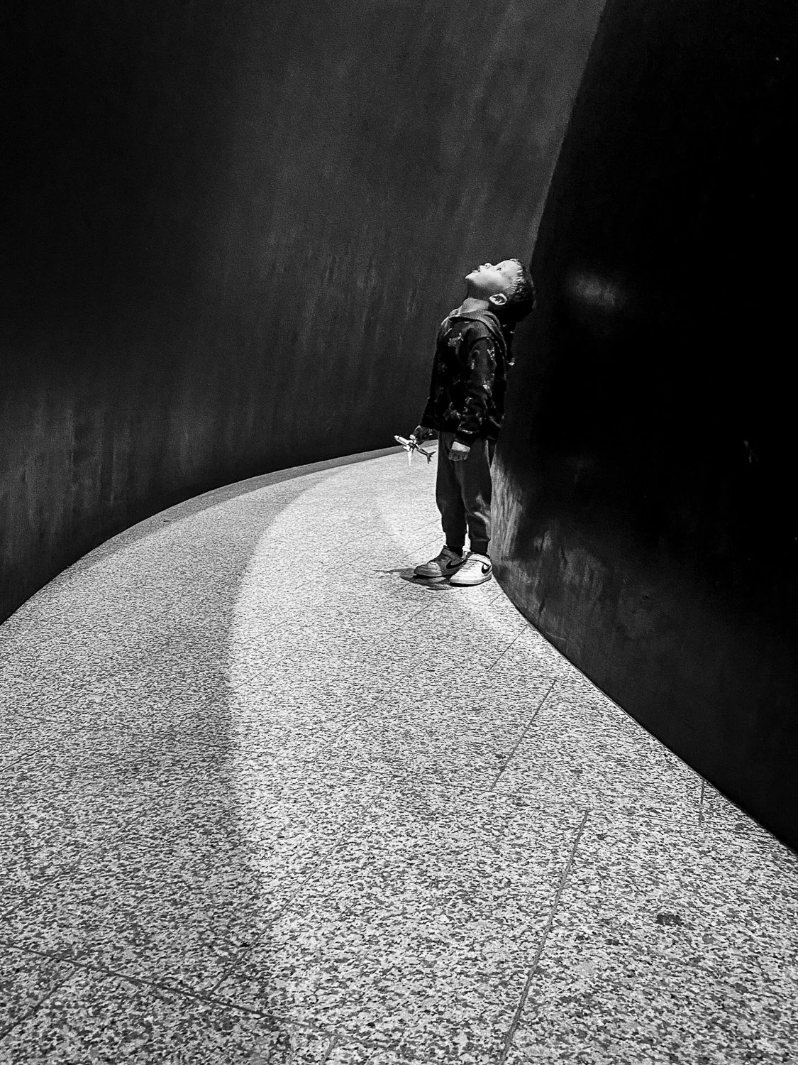 A young boy standing on a curved, textured floor against a dark wall, looking upward, holding a small object in his right hand, in black and white.