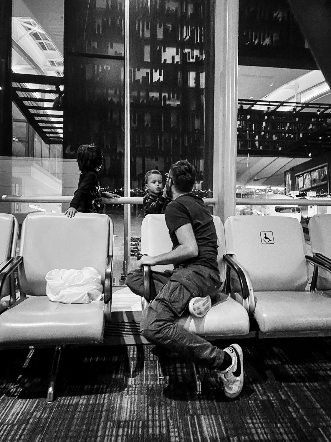 A man with a beard, wearing glasses, a black t-shirt, and cargo pants, is sitting on airport seats and talking to a young girl through a glass window. The girl is standing behind a railing inside the terminal, looking at the man. There are empty airp