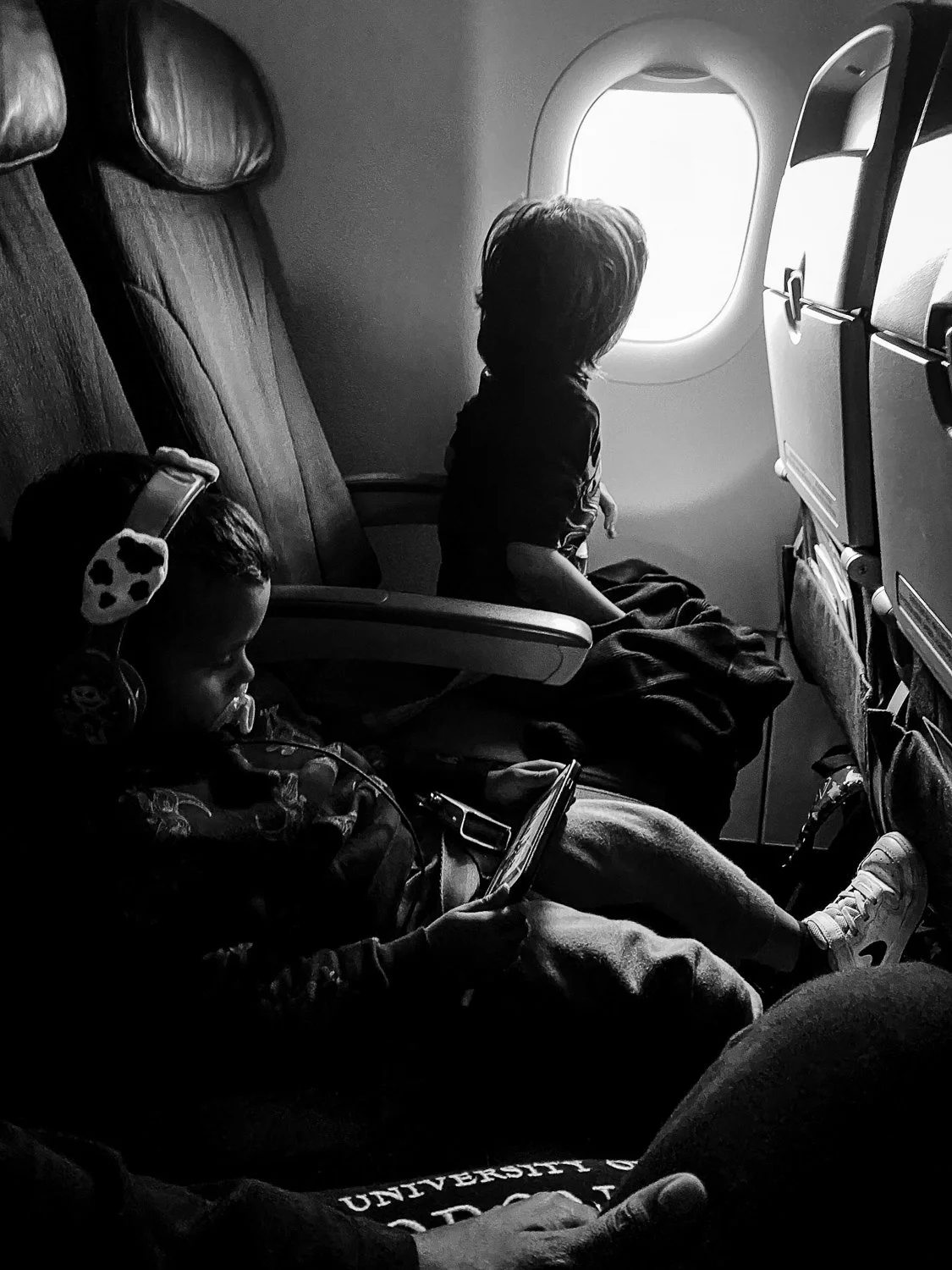 Two children seated on airplane seats, one looking out the window and the other using a tablet, in black and white.