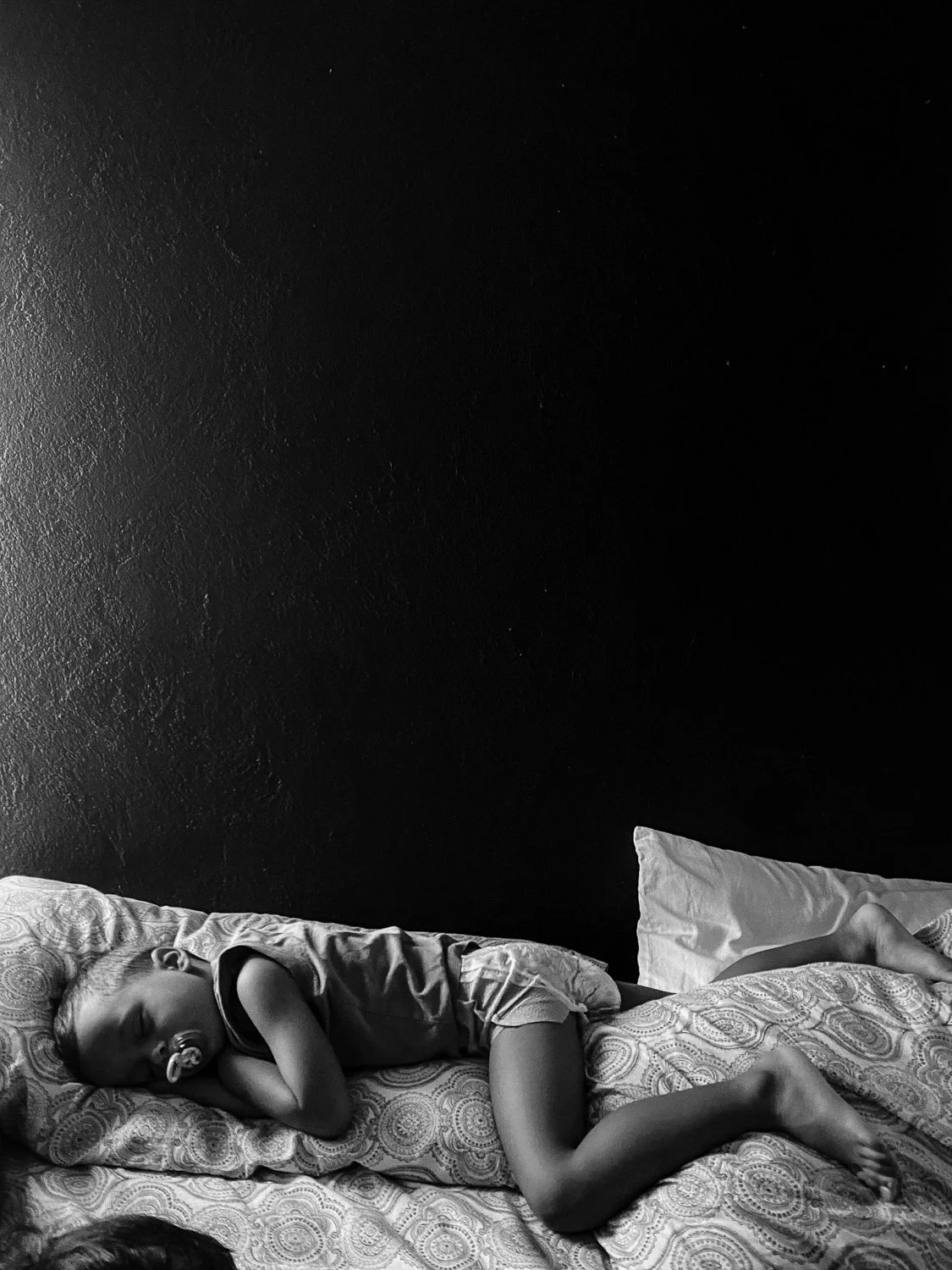 A young child sleeping peacefully on a bed, lying on their side with a pacifier in their mouth, covered with a patterned blanket, and resting against a pillow.