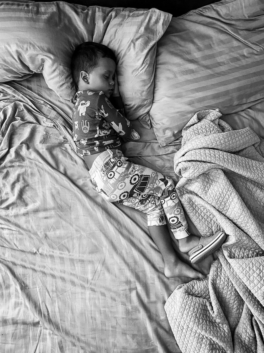 A young boy is sleeping on a bed, curled up on his side with a pillow under his head. He is wearing patterned pajamas with vehicles and is partially covered with a quilt.