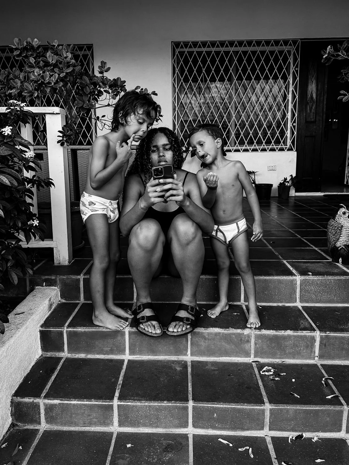 A woman sitting on stairs with two young children, all focusing on a smartphone. The children are shirtless and barefoot, and there is a garden area with plants and a decorative owl on the tiles.
