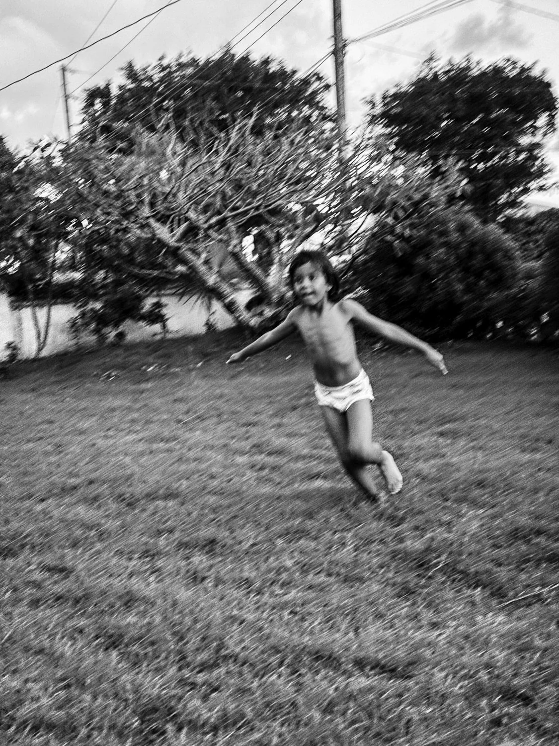 A young child running joyfully in a grassy outdoor area with trees and power lines in the background.