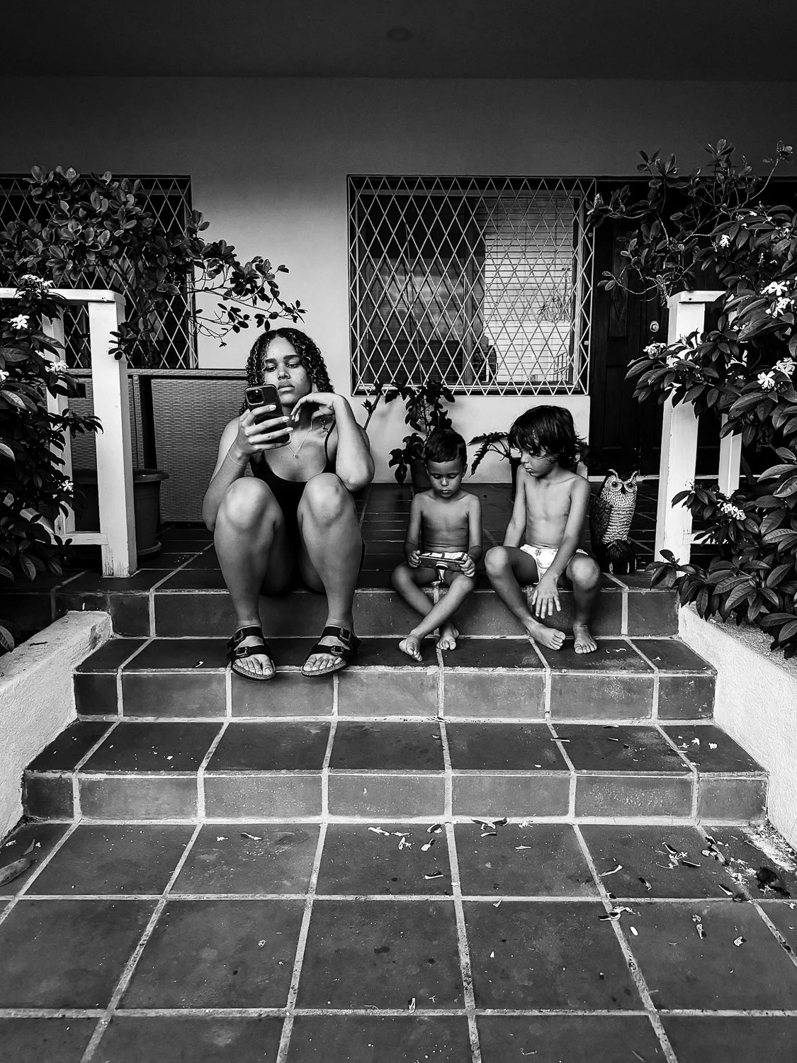 A woman and two children sitting on tiled steps outside, two of the children are looking at handheld devices, and the woman is looking at her phone.