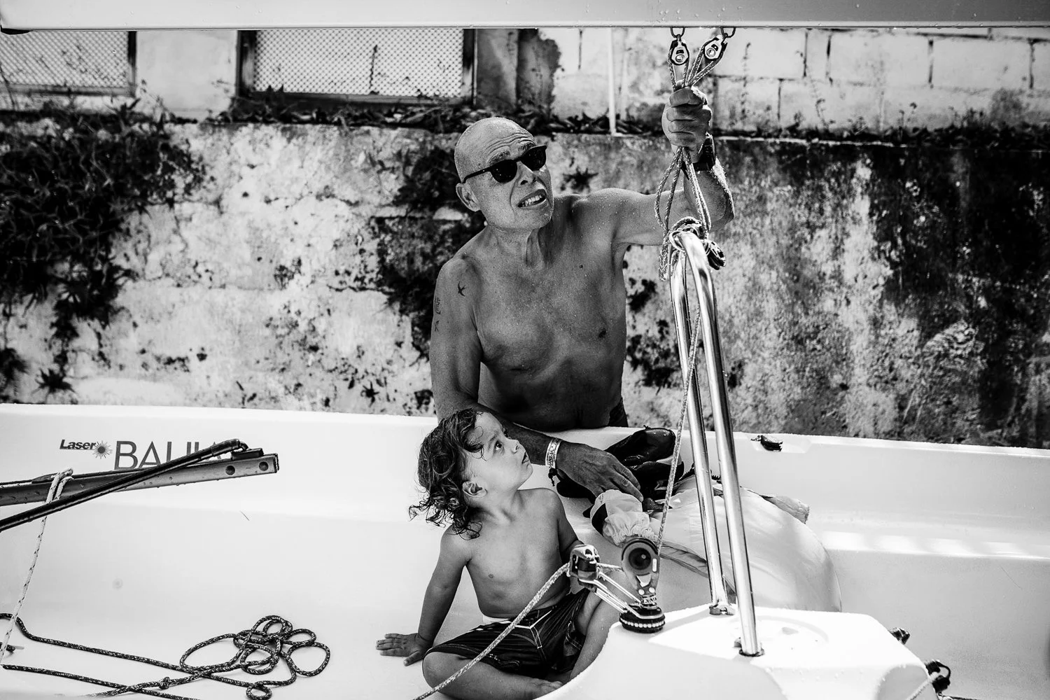 An elderly man with sunglasses and a tattoo on his arm, shirtless, in a boat with a young child. The man is holding ropes, and the child sits on the boat's edge looking up at him. The background shows a stone wall.