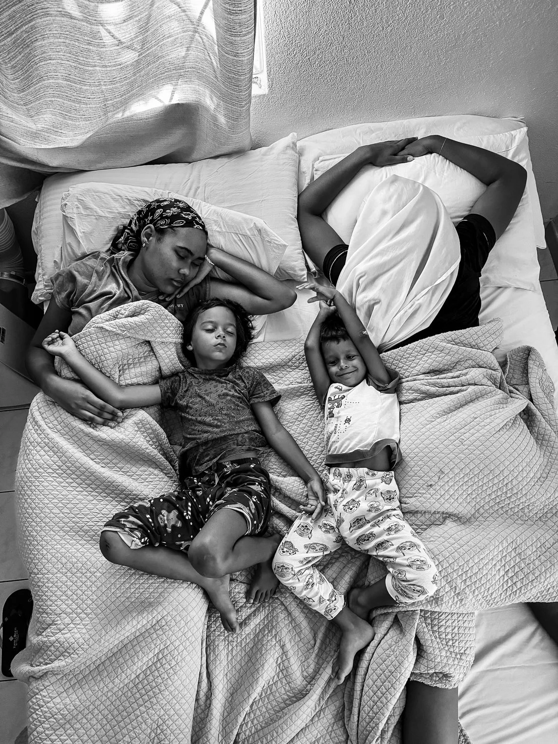 Four children are sleeping on a bed, two of them are holding hands, in a grayscale photo.
