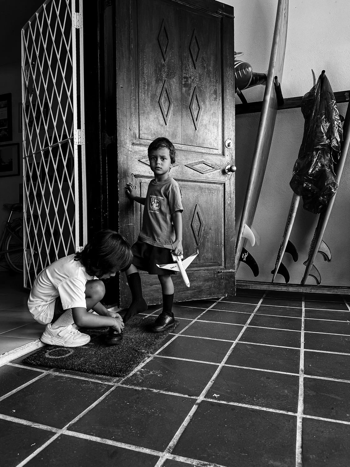Two young children, a boy and a girl, near an open door, with the girl helping the boy put on shoes on a tiled floor in a home.