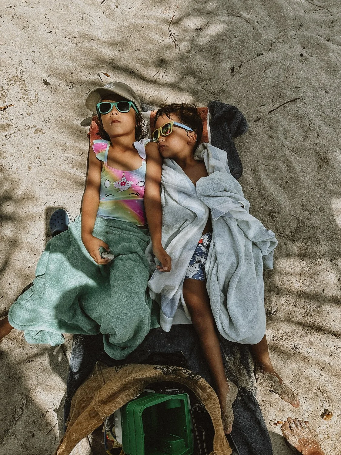Two children with sunglasses and towels lying on a beach towel on the sand, resting. One child is wearing a unicorn-patterned swimsuit and a hat, the other in patterned swim shorts and a shirt. There is a beach bag and sand on their feet.