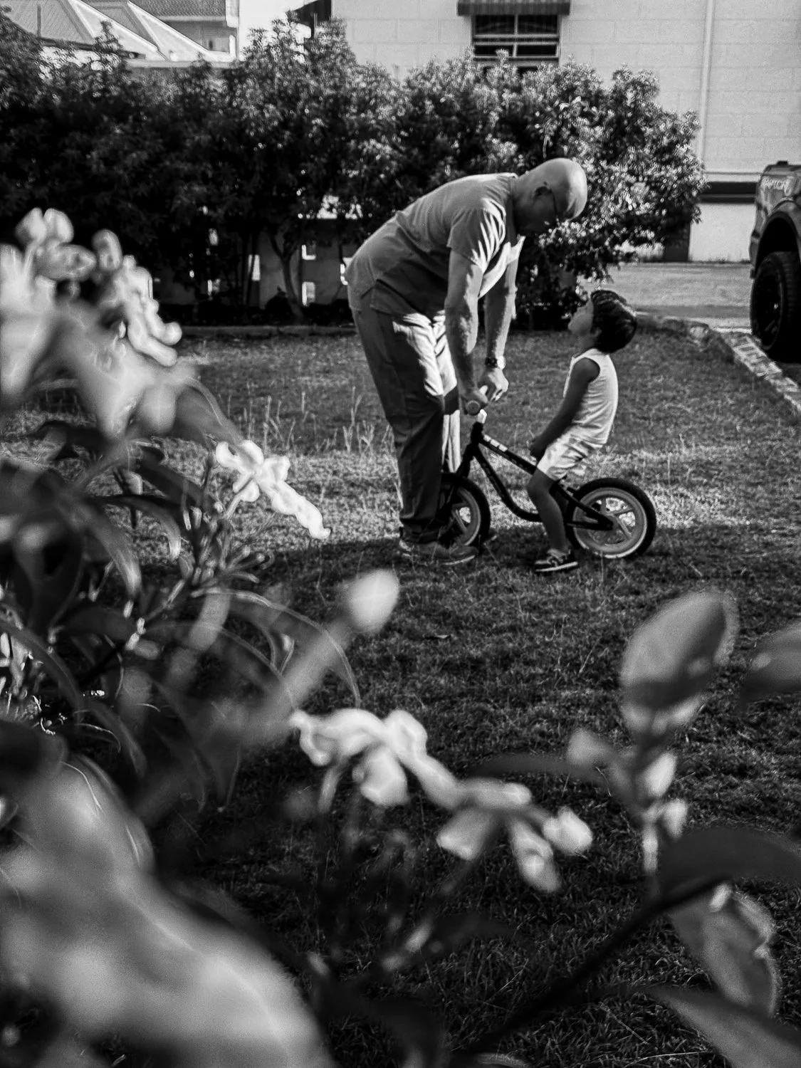 A man helping a young girl learn to ride a bicycle in a backyard on a sunny day, with a bush and house in the background.