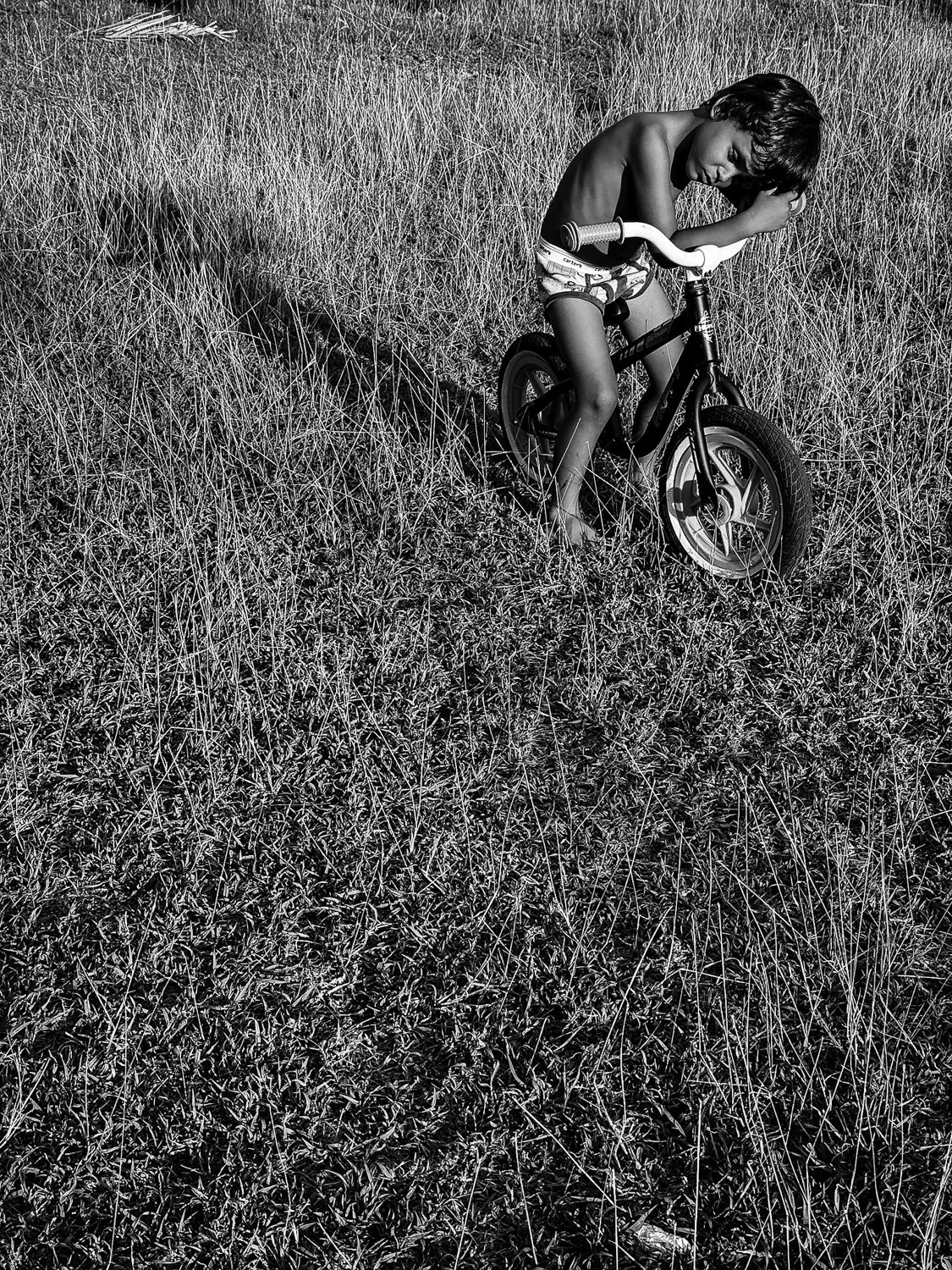 A young boy with curly hair, shirtless and wearing patterned shorts, sitting on a small bicycle in a grassy field, leaning forward with his head resting on his arm.