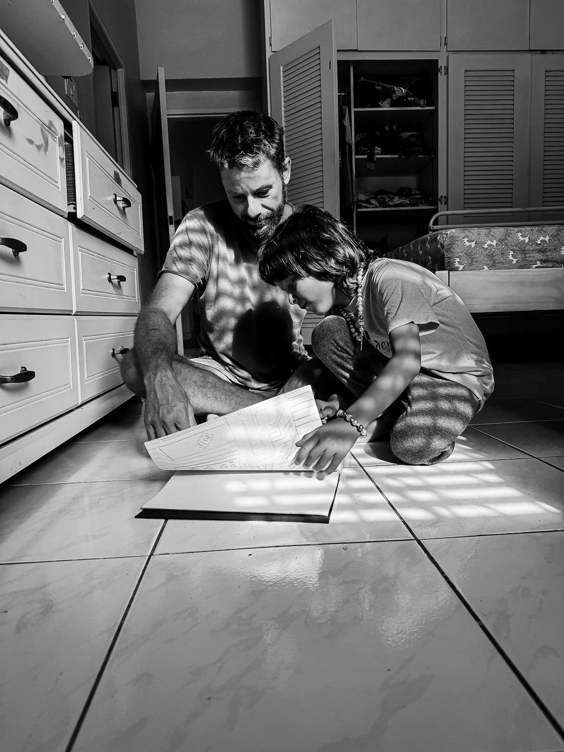 A man and a young girl sitting on the floor of a bedroom, looking at a scrapbook or photo album together. The room has a bed, open closet doors, and cabinets. The image is in black and white.