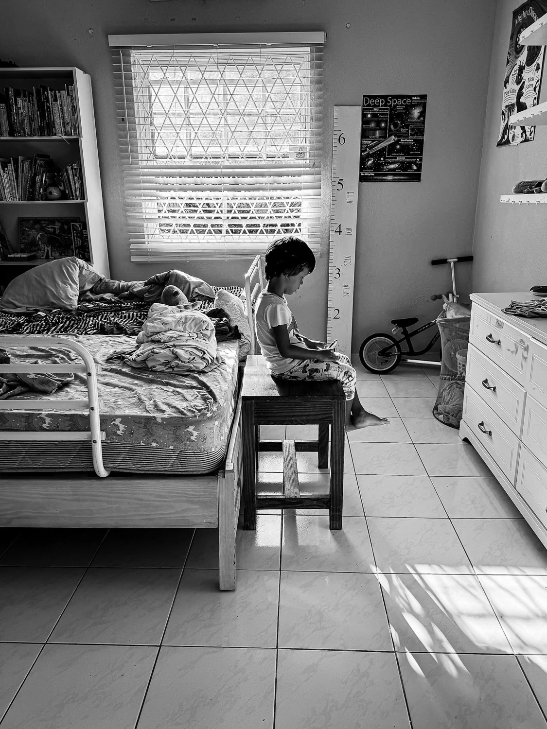 A black and white photo of a child's bedroom with a large window, a bed with disorganized bedding, a bookshelf, and a dresser. A child sits on a small wooden chair near the bed, looking at a device. A balance bike and storage basket are on the floor,