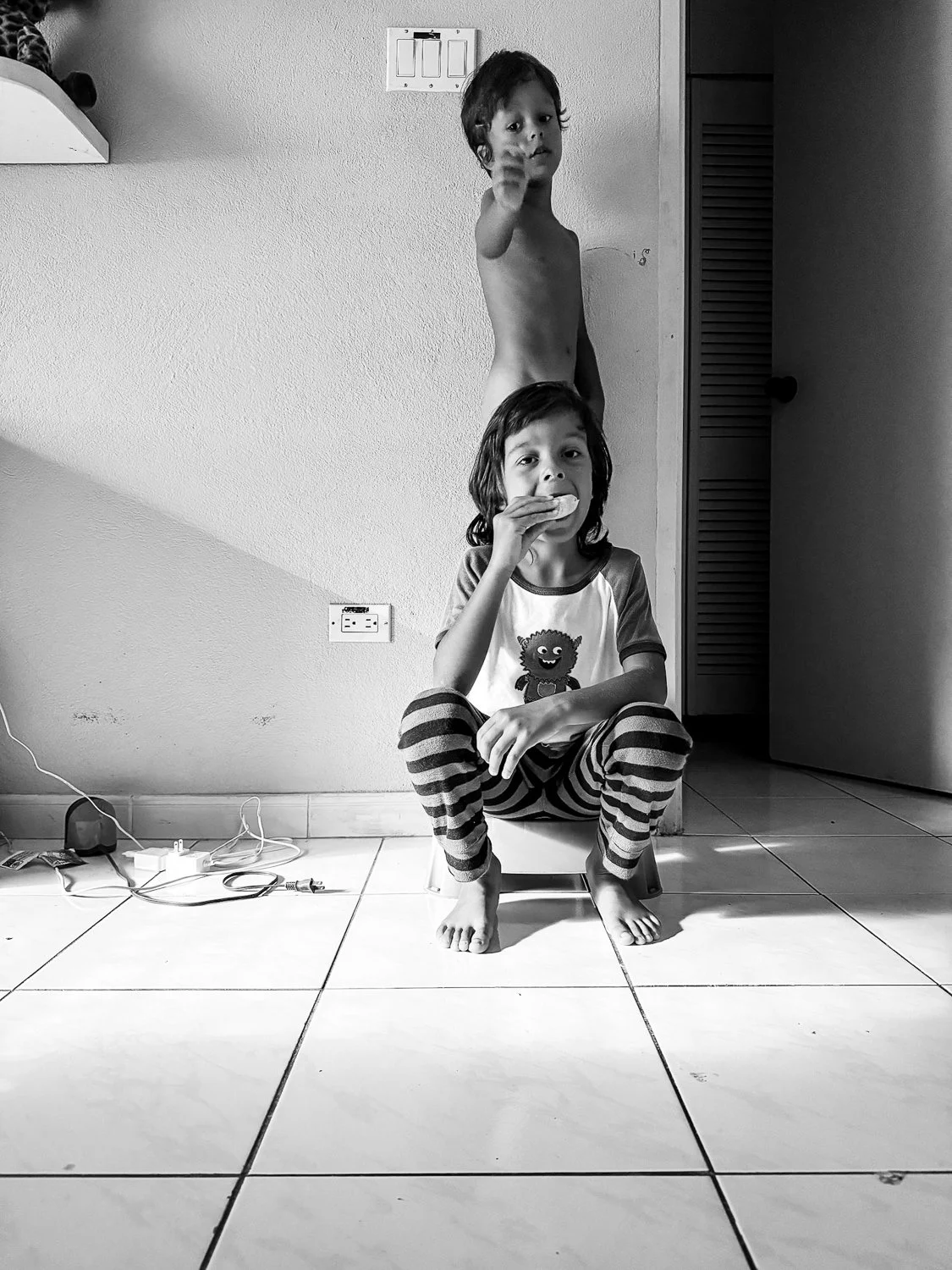 Two young children in a room, one sitting on a small chair eating a snack, the other standing behind, pointing at the camera. The room has a power outlet, a white wall, and a door.
