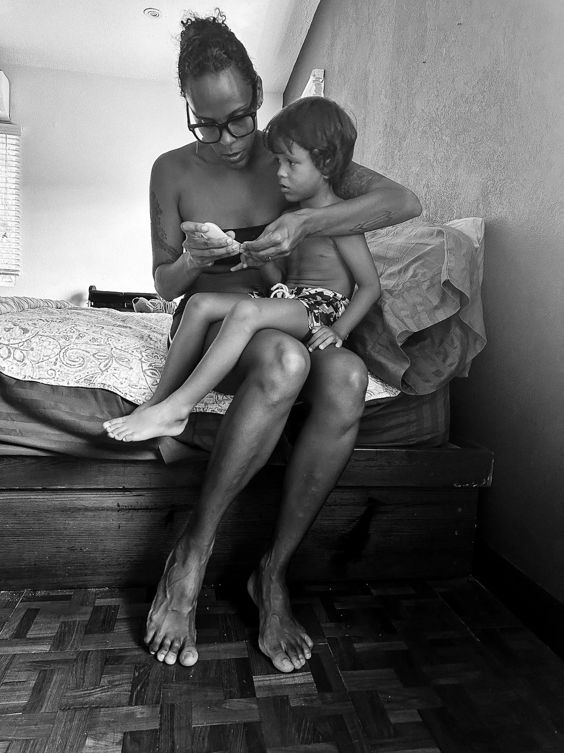 A woman with glasses and tattoos sits on the edge of a bed with a young boy on her lap, both looking at a phone. The woman is topless, and the boy wears shorts. They are in a bedroom with a window, blanket, pillows, and patterned bedspread.