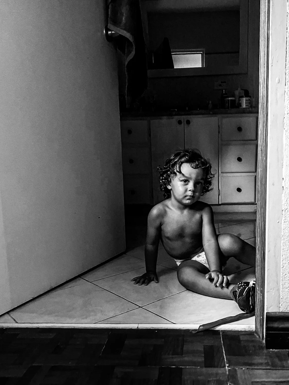 A young shirtless boy with curly hair, wearing a diaper and sneakers, sitting on a tiled floor in a doorway, looking at the camera with a serious expression in a home interior.