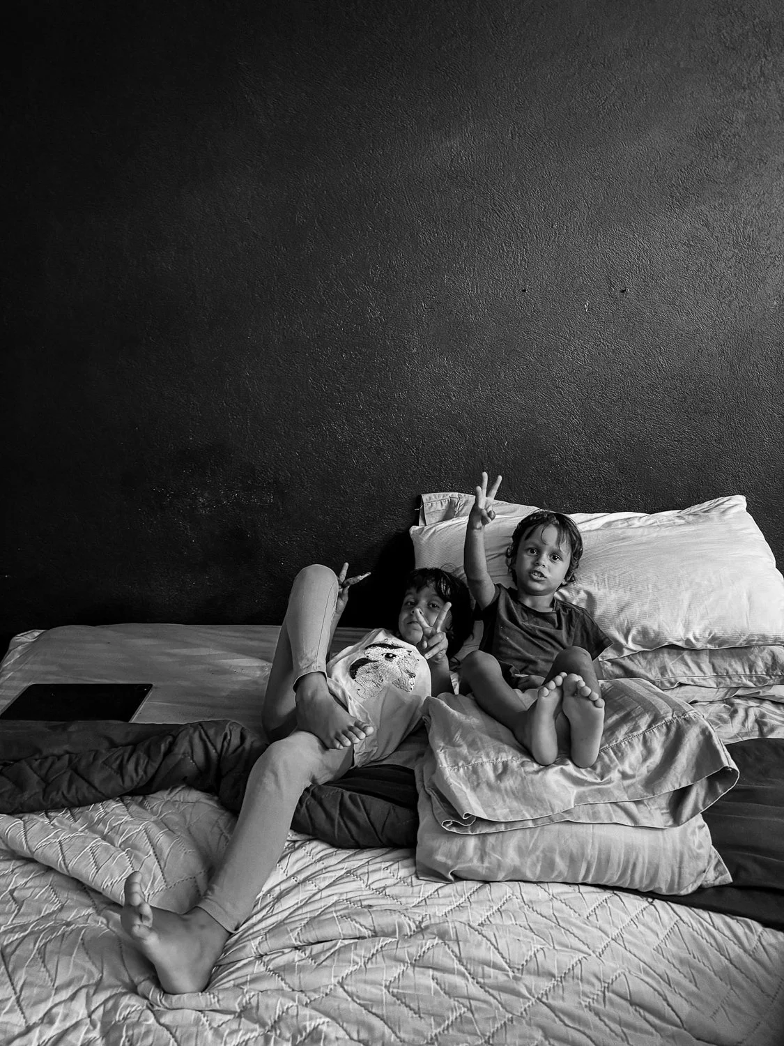 Two children lying on a bed, making peace signs and posing for the camera in a monochrome photo.